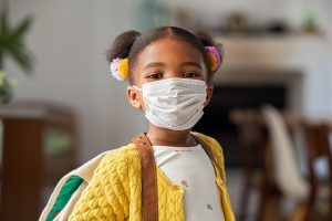 Portrait of black little girl with buns hair wearing bagpack and covid-19 protective face mask ready for elementary school. African american female child wearing surgical mask and looking at camera with school bag. Blacl little girl going back to primary school after quarantine time from corona virus pandemic.